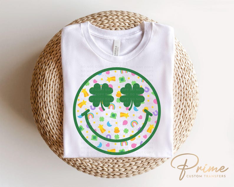 May include: White t-shirt featuring a St. Patrick's Day smiley face design. The smiley face has shamrock eyes and a pattern of rainbows, pots of gold, and shamrocks. The shirt is made of a soft material.