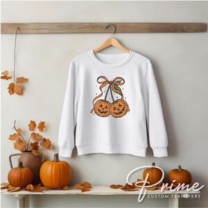 May include: White sweatshirt featuring a Halloween-themed design of two jack-o'-lanterns resembling cherries, tied together with an orange bow and green leaf. The design is centered on the front of the long-sleeved top.