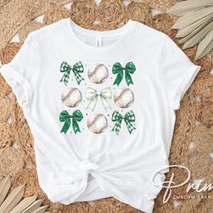 May include: White t-shirt with a green, white, and brown baseball and bow design. The shirt has a knot in the front.