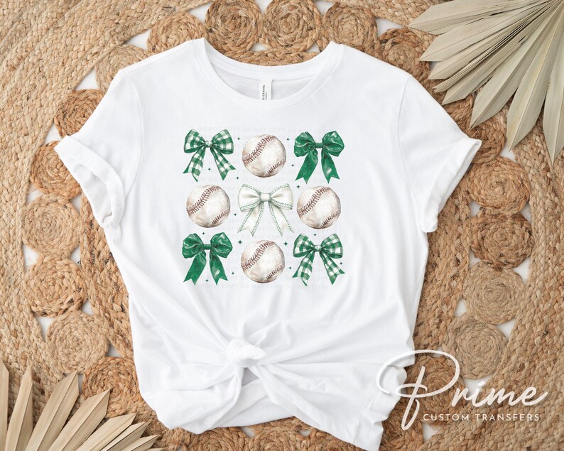 May include: White t-shirt with a green, white, and brown baseball and bow design. The shirt has a knot in the front.