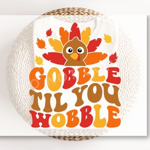 May include: A white t-shirt with a Thanksgiving-themed design. The design features a cartoon turkey with the text "Gobble Til You Wobble" in a retro font.