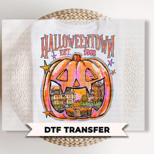 May include: A white t-shirt featuring a graphic print with the text HALLOWEENTOWN EST. 1998. The design includes a large orange jack-o'-lantern with a detailed illustration of a house and landscape inside, accented by small colorful stars.