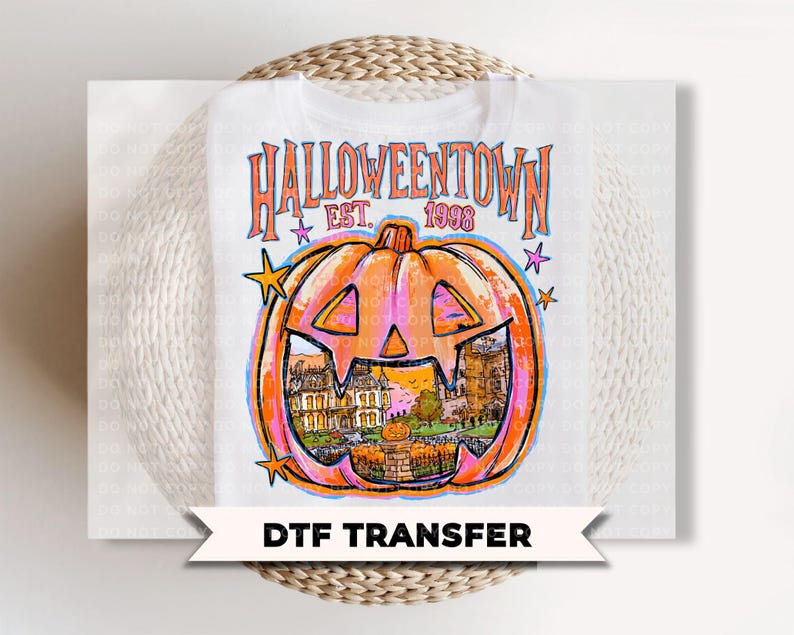 May include: A white t-shirt featuring a graphic print with the text HALLOWEENTOWN EST. 1998. The design includes a large orange jack-o'-lantern with a detailed illustration of a house and landscape inside, accented by small colorful stars.