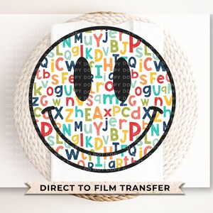 May include: A black and white smiley face with a colorful background of letters. The letters are in a variety of colors, including red, orange, yellow, green, blue, and purple. The smiley face is a simple design with a black outline and a black smile.