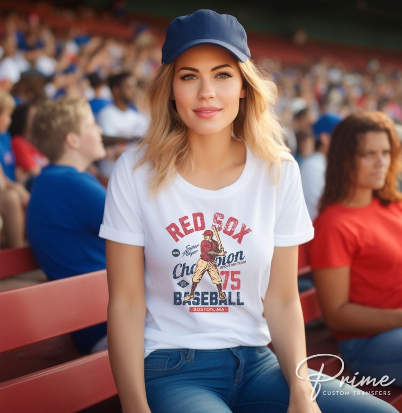 Może przedstawiać: Biała koszulka z vintage'ową grafiką baseballową przedstawiającą gracza w czerwonym stroju z napisem "Red Sox" w czerwono-niebieskich literach. Grafika zawiera r&oacute;wnież tekst "Super Player Champion Sporting Team 75 Baseball Boston, MA".