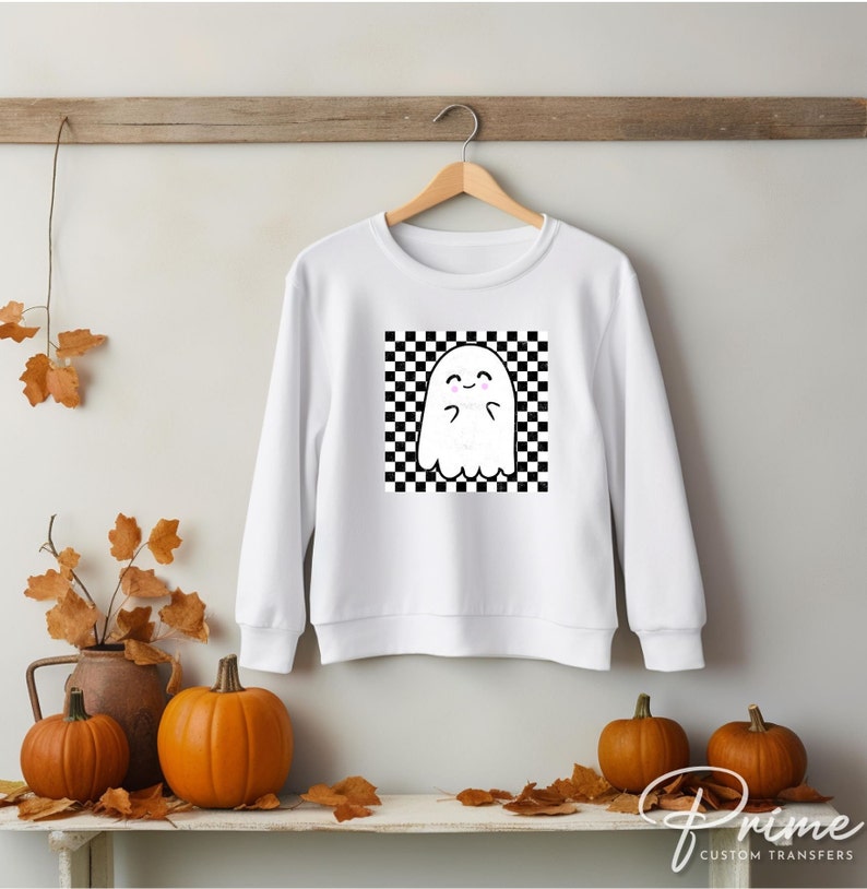 May include: White sweatshirt with a black and white checkered square design featuring a cartoon ghost with a smiling face.