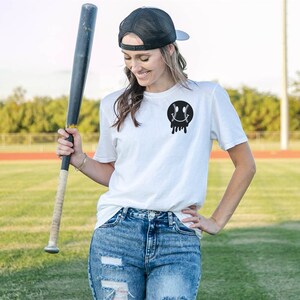 May include: A white t-shirt featuring a black baseball-shaped smiley face graphic. The shirt is being worn by a person holding a wooden baseball bat.