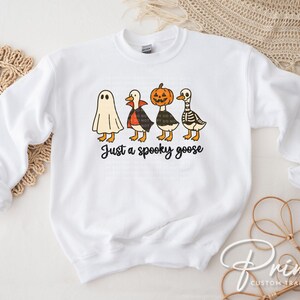 May include: White sweatshirt featuring a Halloween-themed graphic. The design includes four cartoon geese dressed as a ghost, vampire, pumpkin, and skeleton. The phrase "Just a spooky goose" is written in black script below the geese.
