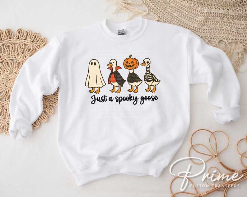 May include: White sweatshirt featuring a Halloween-themed graphic. The design includes four cartoon geese dressed as a ghost, vampire, pumpkin, and skeleton. The phrase "Just a spooky goose" is written in black script below the geese.