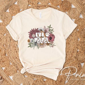 May include: Cream-colored t-shirt with the text "FUR MAMA" in a floral design. The design includes a paw print and various flowers in shades of pink, white, and green. The shirt is displayed on a woven, circular rug.