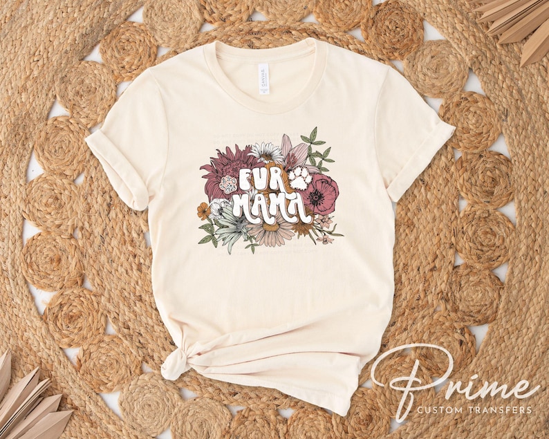 May include: Cream-colored t-shirt with the text "FUR MAMA" in a floral design. The design includes a paw print and various flowers in shades of pink, white, and green. The shirt is displayed on a woven, circular rug.