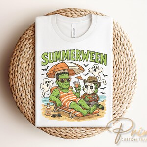 May include: White t-shirt with a graphic of Frankenstein and Freddy Krueger relaxing on a beach under an orange and tan umbrella. The text "Summerween" is above the characters. The shirt is folded on a woven, circular surface.