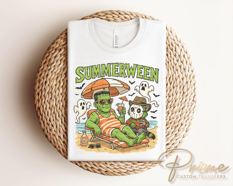 May include: White t-shirt with a graphic of Frankenstein and Freddy Krueger relaxing on a beach under an orange and tan umbrella. The text "Summerween" is above the characters. The shirt is folded on a woven, circular surface.