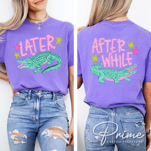 May include: A purple t-shirt with a front graphic that says "LATER" above a teal alligator illustration. The back of the shirt says "AFTER WHILE" above a matching alligator. Pink and yellow star accents are included.