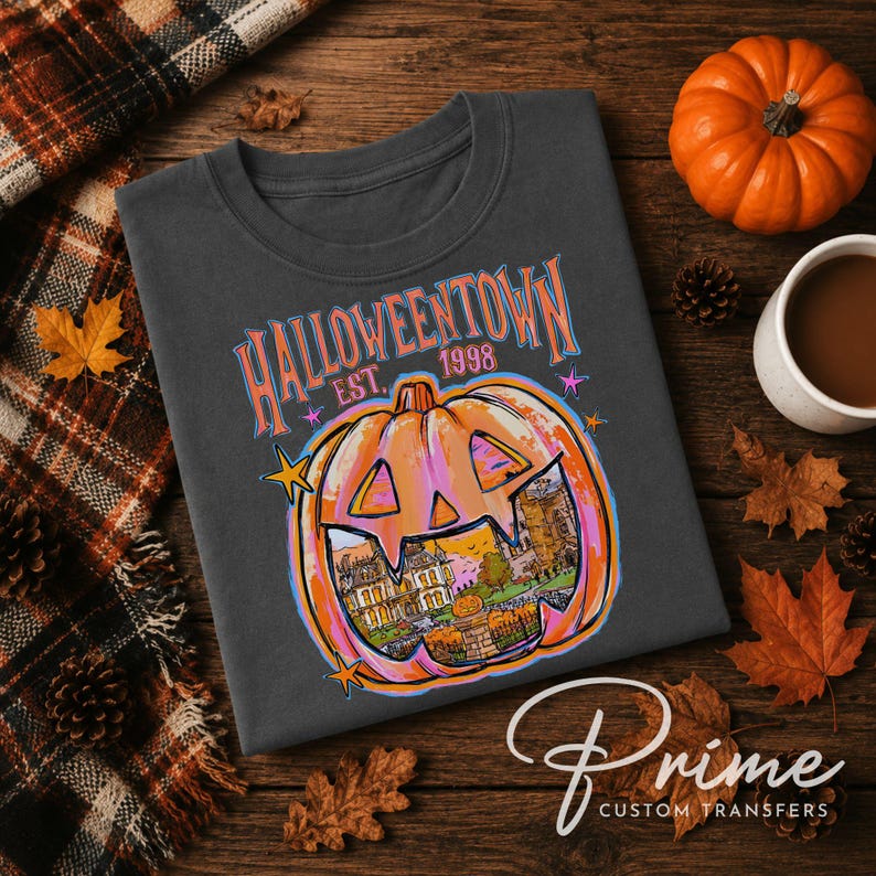 May include: A dark gray t-shirt featuring a graphic of a carved pumpkin with a castle scene inside. The text reads HALLOWEENTOWN EST. 1998 in orange and blue letters. The design includes small star accents and a vibrant orange and pink color palette.