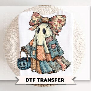May include: A white t-shirt featuring a graphic of a ghost made of patchwork fabric in shades of blue, orange, and tan. The ghost wears a large floral bow and carries a blue jack-o'-lantern bucket. The text DTF TRANSFER is printed below.