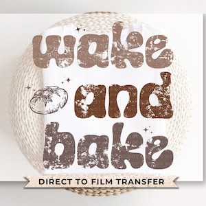 May include: A white kitchen towel with a brown distressed graphic that says "Wake and Bake" with a small illustration of a loaf of bread. The text "Direct to Film Transfer" is printed below the graphic.