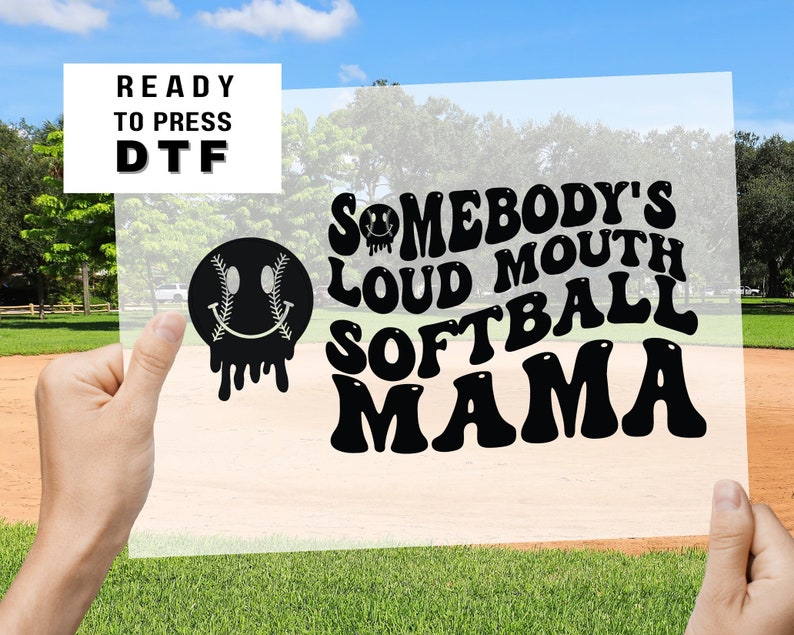 May include: Black and white graphic design featuring the text "Somebody's Loud Mouth Softball Mama" in a retro style font. A black and white smiley face with a baseball seam design is also included.