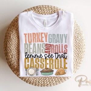 May include: White t-shirt with a Thanksgiving themed graphic that says "Turkey Gravy Beans and Rolls lemme see that Casserole".