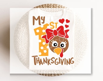 Op de afbeelding: Witte romper met de tekst "My 1st Thanksgiving" in bruine letters. Het cijfer 1 is geel met witte stippen en een rode strik. Een kalkoen gezicht met een rode strik bevindt zich bovenop het cijfer 1.
