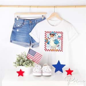 May include: White t-shirt with a red, white, and blue goose wearing sunglasses and holding a hot dog. The goose is surrounded by fireworks and a checkered background. The text "Prime" is visible in the bottom right corner.