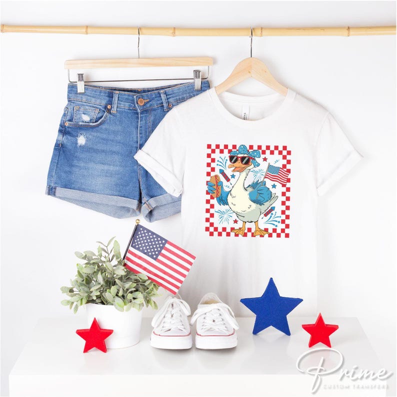 May include: White t-shirt with a red, white, and blue goose wearing sunglasses and holding a hot dog. The goose is surrounded by fireworks and a checkered background. The text "Prime" is visible in the bottom right corner.