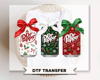 May include: A white t-shirt featuring a DTF transfer design of three soda cans labeled Dr Pepper. The cans are decorated with Christmas patterns, including ornaments, snowflakes, and gingerbread men, and each is topped with a large fabric bow.