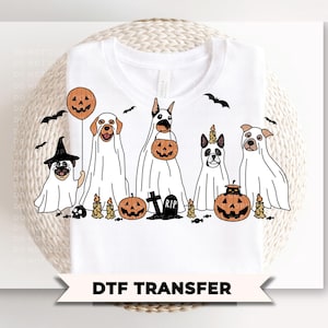 May include: A white t-shirt featuring a graphic of five dogs dressed as ghosts for Halloween. The dogs are surrounded by orange jack-o'-lanterns, candles, bats, and a tombstone with the text RIP. The design includes a balloon and candy.