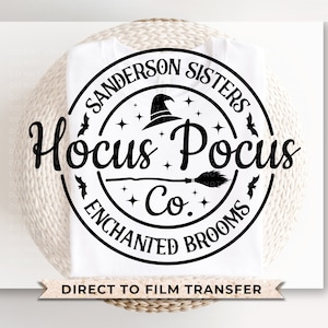 May include: Black and white graphic design of a circle with the text "Sanderson Sisters Hocus Pocus Co. Enchanted Brooms" and a witch's hat and broom inside the circle. The graphic is on a white t-shirt.