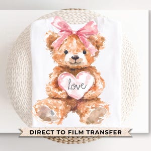 May include: A white t-shirt with a watercolor illustration of a brown teddy bear wearing a pink bow and holding a pink heart that says "Love".