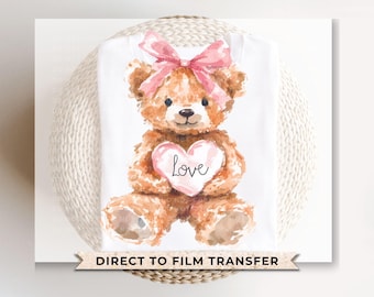 Peut inclure: Un t-shirt blanc avec une illustration aquarelle d'un ours en peluche brun portant un nœud rose et tenant un cœur rose qui dit "Love".