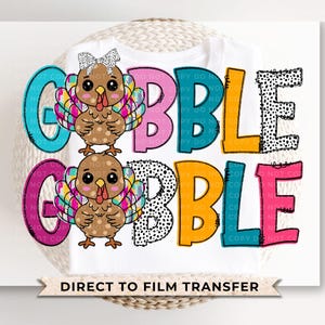 May include: A white t-shirt with a colorful "Gobble" design featuring two cartoon turkeys with pink, blue, yellow, and black accents. The text "Direct to Film Transfer" is printed at the bottom of the shirt.