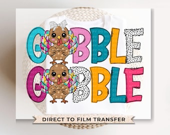 May include: A white t-shirt with a colorful "Gobble" design featuring two cartoon turkeys with pink, blue, yellow, and black accents. The text "Direct to Film Transfer" is printed at the bottom of the shirt.