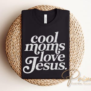 May include: Black t-shirt with the text "cool moms love Jesus" in white, displayed on a woven, circular surface. The shirt is made of a soft material and is suitable for casual wear.