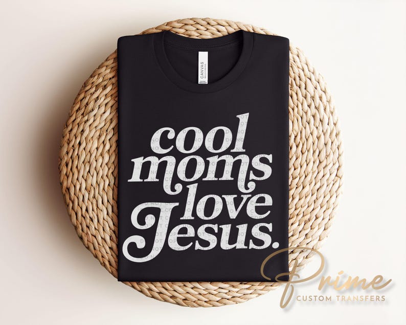 May include: Black t-shirt with the text "cool moms love Jesus" in white, displayed on a woven, circular surface. The shirt is made of a soft material and is suitable for casual wear.