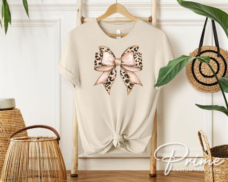May include: A beige t-shirt with a large pink and brown leopard print bow on the front.