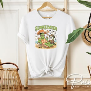 May include: White t-shirt featuring a colorful graphic of Frankenstein and Freddy Krueger relaxing on a beach under an umbrella. The text "SUMMERWEEN" is above the graphic. The shirt is hanging on a wooden hanger.