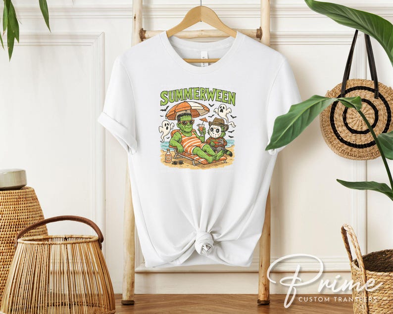May include: White t-shirt featuring a colorful graphic of Frankenstein and Freddy Krueger relaxing on a beach under an umbrella. The text "SUMMERWEEN" is above the graphic. The shirt is hanging on a wooden hanger.