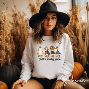 May include: White sweatshirt with a Halloween-themed graphic of four cartoon geese dressed as a ghost, vampire, pumpkin, and skeleton. The text "Just a spooky goose" is below the geese. The sweatshirt is worn by a person wearing a black hat.