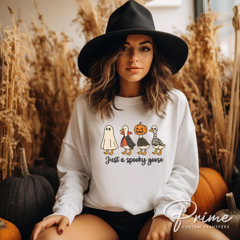 May include: White sweatshirt with a Halloween-themed graphic of four cartoon geese dressed as a ghost, vampire, pumpkin, and skeleton. The text "Just a spooky goose" is below the geese. The sweatshirt is worn by a person wearing a black hat.