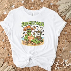 May include: White t-shirt with a colorful graphic that reads "Summerween." The graphic features Frankenstein and Freddy Krueger relaxing on a beach, under an umbrella. The design includes ghosts, a beach scene, and a cocktail.