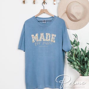Puede incluir: Una camiseta azul claro con las palabras "MADE FOR MORE" en un dise&ntilde;o floral. El texto "PHILIPPIANS 1:1" est&aacute; debajo del texto principal. La camiseta est&aacute; colgada en una percha de madera.