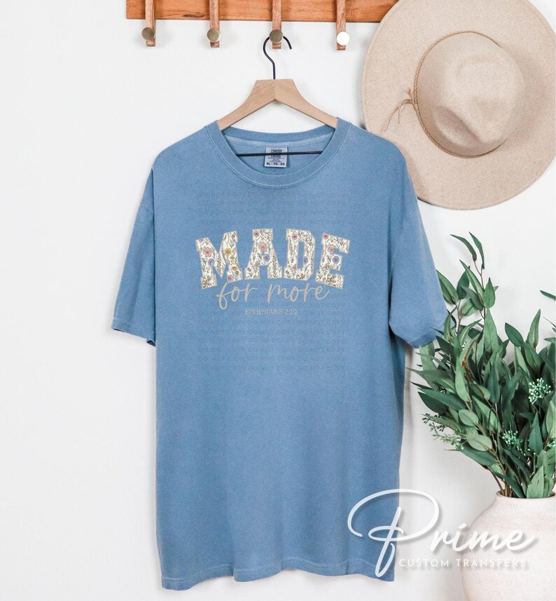 Puede incluir: Una camiseta azul claro con las palabras "MADE FOR MORE" en un dise&ntilde;o floral. El texto "PHILIPPIANS 1:1" est&aacute; debajo del texto principal. La camiseta est&aacute; colgada en una percha de madera.