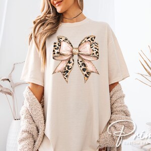 May include: A beige t-shirt with a large, pink and brown leopard print bow on the front. The bow has a glittery texture.