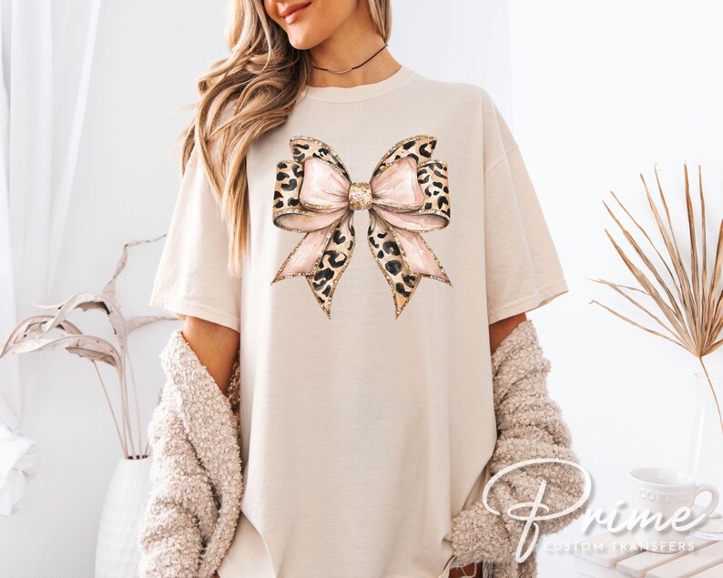May include: A beige t-shirt with a large, pink and brown leopard print bow on the front. The bow has a glittery texture.