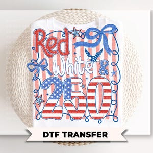 May include: A white t-shirt featuring a graphic design with the text Red White & 250. The design includes red and white vertical stripes, blue stars, a blue eagle, and blue ribbon accents. The numbers 250 are filled with a stars and stripes pattern.