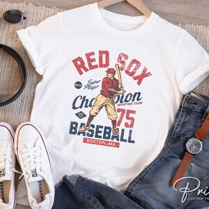 Może przedstawiać: Biała koszulka z grafiką w stylu vintage przedstawiającą baseballistę w czerwonym stroju z napisem "Red Sox" w kolorze czerwonym i niebieskim, "Super Player" w kolorze czarnym, "Champion" w kolorze niebieskim, "Sporting Team" w kolorze czarnym, "75" w kolorze czerwonym i "Baseball" w kolorze niebieskim. Tekst "Boston, MA" jest w kolorze czarnym.