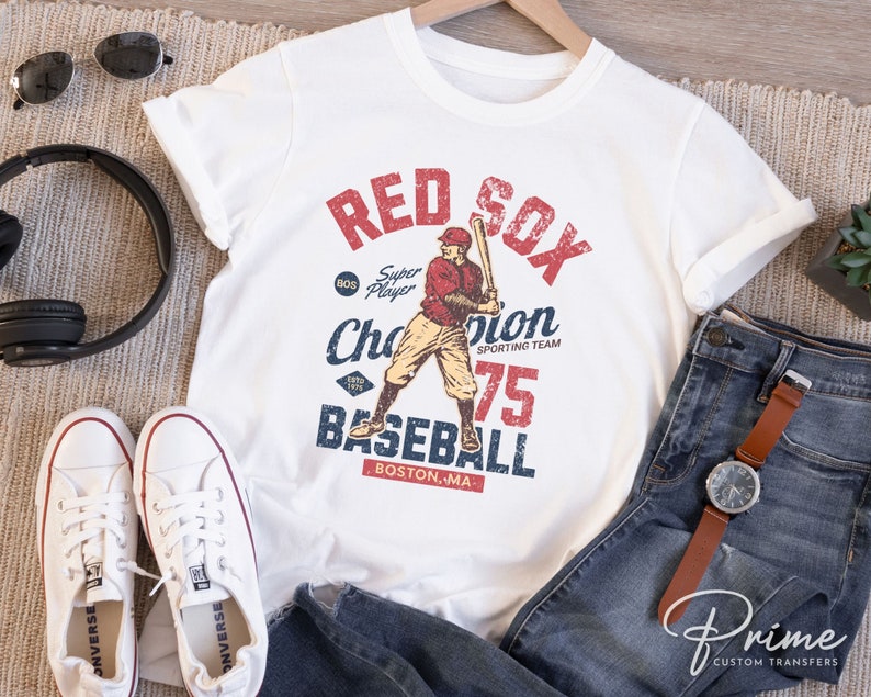 Może przedstawiać: Biała koszulka z grafiką w stylu vintage przedstawiającą baseballistę w czerwonym stroju z napisem "Red Sox" w kolorze czerwonym i niebieskim, "Super Player" w kolorze czarnym, "Champion" w kolorze niebieskim, "Sporting Team" w kolorze czarnym, "75" w kolorze czerwonym i "Baseball" w kolorze niebieskim. Tekst "Boston, MA" jest w kolorze czarnym.