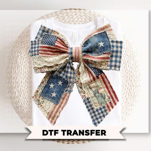 May include: White t-shirt with a large patriotic bow design. The bow features sections of the American flag, blue gingham, and floral lace. The bow is centered on the shirt, with the text "DTF TRANSFER" below.