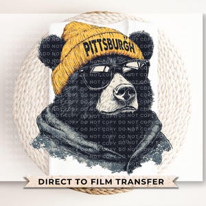 May include: A graphic of a black bear wearing a yellow beanie with "PITTSBURGH" printed on it, black sunglasses, and a gray hoodie. The image is a direct-to-film transfer.